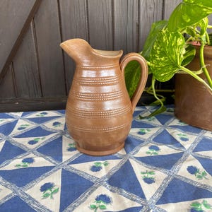 May include: A brown ceramic pitcher with a textured surface and a handle. The pitcher is sitting on a blue and white patterned tablecloth.