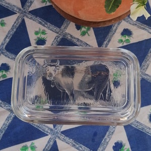 May include: A clear glass butter dish with a cow design. The rectangular dish has a lid and features an embossed image of a cow. The dish sits on a blue and white patterned tablecloth, with a terracotta pot of flowers in the background.