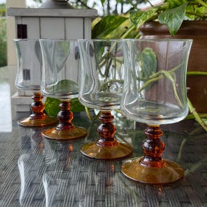 Può includere: Quattro calici di vetro trasparente con steli e basi color ambra sono disposti su un tavolo di vetro. I calici hanno un design curvo unico. Lo sfondo comprende vegetazione e una piccola casa decorativa bianca.