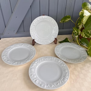 Può includere: Quattro piatti in ceramica bianca con disegni floreali e di frutta in rilievo sui bordi. Un piatto è esposto su un supporto di legno. I piatti sono disposti su una tovaglia chiara, con una porta di legno grigia sullo sfondo.