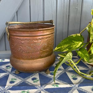Könnte beinhalten: Ein Vintage-Kupfereimer mit Griff und dekorativer floraler Ätzung. Der Eimer steht auf drei kleinen Füßen und hat einen bronzenen Griff. Der Eimer wird auf einer blau-weiß gemusterten Tischdecke präsentiert.