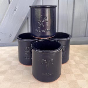 Peut inclure: Un ensemble de quatre tasses en céramique bleu marine foncé disposées en groupe. Chaque tasse présente un symbole du zodiaque en creux et son nom correspondant, dont Sagittaire, Cancer et Bélier, avec une finition lisse et une base en terre cuite.