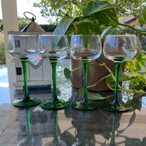 Op de afbeelding: Vier heldere wijnglazen met groene stelen en voetjes staan op een glazen tafel. De glazen worden weerspiegeld in het tafeloppervlak. Op de achtergrond staat een potplant met groene bladeren.