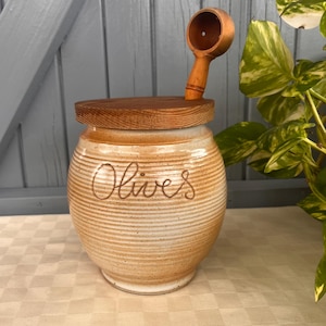 May include: A ceramic olive jar with a wooden lid and matching scoop. The jar is tan with the word "Olives" in cursive. The lid and scoop are made of light brown wood. The jar is sitting on a checkered tablecloth.