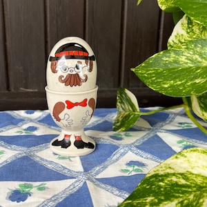 May include: A white ceramic egg cup with a cartoon face and a red bow tie. The egg cup is holding a white egg with a cartoon face, black hat, and red and black glasses. The egg cup is sitting on a blue and white checkered tablecloth.