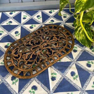May include: An oval, bronze-coloured trivet with an ornate, openwork design. The trivet features a detailed floral and vine pattern. It is placed on a blue and white patterned tablecloth with a floral motif, suitable for kitchen or dining decor.
