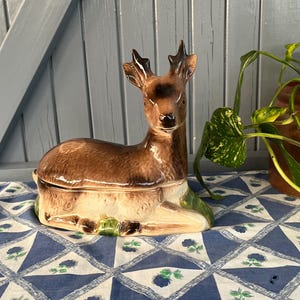 Może przedstawiać: Ceramiczna figurka jelenia z brązowym korpusem, kremowym brzuchem i ciemnobrązowymi rogami. Jeleń leży na niebiesko-białej wzorzystej tkaninie. Figurka ma pokrywkę i prawdopodobnie jest pojemnikiem dekoracyjnym.