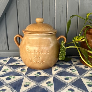 Può includere: Contenitore in ceramica beige con coperchio e due manici. Le parole "Poubelle de table" sono impresse sul davanti. Il contenitore è appoggiato su una tovaglia a motivi blu e bianchi.