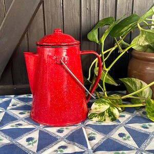 Op de afbeelding: Rode geëmailleerde camping koffiepot met een metalen handvat. De pot staat op een blauw-wit geblokt tafelkleed.
