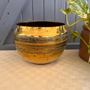 Pode incluir: Uma taça de latão polido com um corpo largo e arredondado, apresentando duas faixas horizontais com padrões intrincados de flores e volutas em relevo. A superfície metálica tem um tom dourado quente e reflexivo com variações sutis.