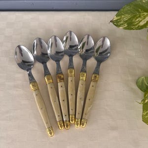 May include: A set of six silver-toned metal spoons with cream-colored, marbled resin handles. Each spoon features gold-toned metal accents at the top and bottom of the handle, with a smooth, polished finish on the bowl and a decorative rivet detail.