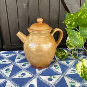 May include: A brown ceramic teapot with a lid and a handle. The teapot is sitting on a blue and white tablecloth with a floral pattern.