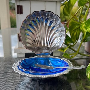 Puede incluir: Un plato decorativo en forma de concha con exterior plateado e interior azul vibrante. El plato está abierto, revelando una pequeña cuchara. El diseño recuerda a una concha, perfecto para servir pequeñas porciones o como acento decorativo.