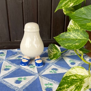 Puede incluir: Un salero y pimentero de cerámica blanca con forma de persona con zapatos azules y medias con estampado naranja y blanco. El salero está sobre un mantel con estampado azul y blanco.