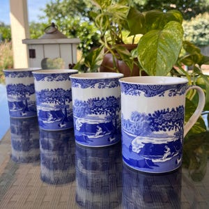 Puede incluir: Cuatro tazas de cerámica blanca con un diseño de escena pastoral azul. Las tazas tienen un asa y una forma cilíndrica. El diseño azul representa un paisaje con figuras y árboles. Las tazas se exhiben sobre una superficie reflectante.