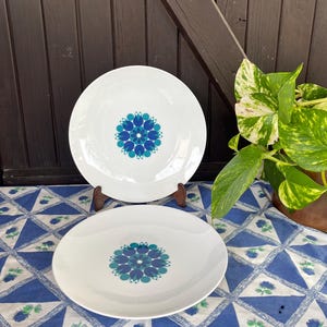 Op de afbeelding: Twee witte keramische dinerborden met een blauw en groen bloemenontwerp. De borden liggen op een blauw en wit tafelkleed.