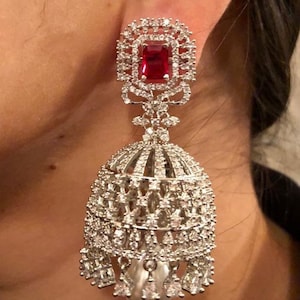 Peut inclure: Boucles d'oreilles pendantes argentées avec une pierre précieuse rouge et une perle pendante. Les boucles d'oreilles sont décorées de détails complexes et de pierres scintillantes.