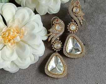 May include: A pair of gold earrings with a bird design and large clear stones. The earrings are set against a gray background with white flowers.