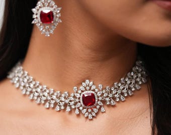 May include: A silver choker necklace featuring a large red gemstone surrounded by smaller white gemstones. The necklace is crafted from a delicate chain with a floral design.