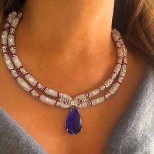 Puede incluir: Un collar de dos hilos con una cadena de color plateado, adornado con pequeñas piedras transparentes y detalles rojos. Una gran piedra azul oscuro en forma de lágrima cuelga del centro. El collar se lleva alrededor del cuello.
