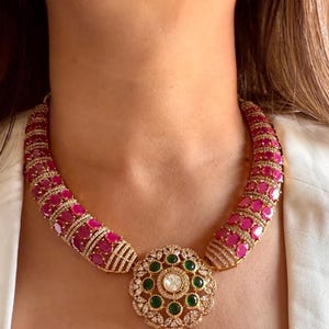 May include: A gold necklace with a large, round pendant featuring a white stone surrounded by green and white stones. The necklace is made of gold and has a row of pink stones on each side.