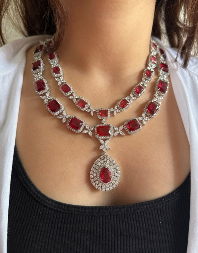 May include: A two-strand necklace featuring large, rectangular, and pear-shaped red gemstones, accented with smaller clear stones. The necklace is set in a silver-colored metal, creating a luxurious and elegant look. The necklace is worn over a black top.
