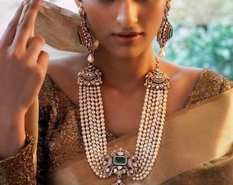 May include: A woman wearing a gold-toned necklace and matching earrings. The necklace features multiple strands of small, white pearls and a large, ornate pendant with a green gemstone. The earrings also have green gemstones and pearl drops.