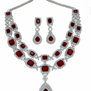 May include: A silver-toned jewelry set featuring a necklace and matching earrings. The necklace has two strands with square and rectangular red gemstones, and a teardrop pendant. The earrings have a similar design with red gemstones.