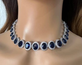 May include: A silver necklace with oval, dark blue gemstones, each encircled by smaller, clear stones. The necklace is displayed on a mannequin. The dark blue stones are evenly spaced along the necklace's length, creating a symmetrical design.