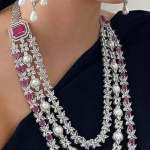 K&ouml;nnte beinhalten: Eine dreireihige silberne Halskette mit rosa und wei&szlig;en Steinen. Die Halskette hat ein florales Design mit kleinen rosa Steinen und gr&ouml;&szlig;eren wei&szlig;en Steinen. Die Halskette ist mit einem gr&ouml;&szlig;eren rosa Stein in der Mitte jeder Reihe verziert.