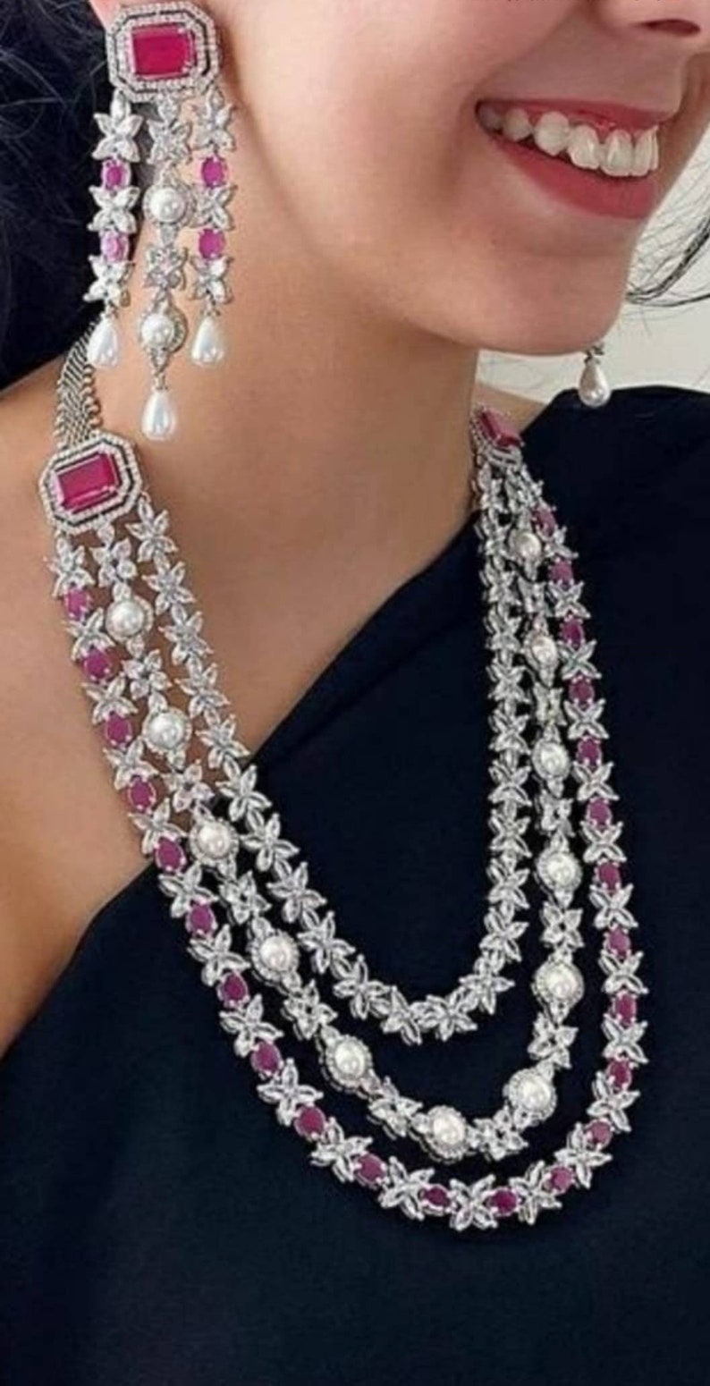 K&ouml;nnte beinhalten: Eine dreireihige silberne Halskette mit rosa und wei&szlig;en Steinen. Die Halskette hat ein florales Design mit kleinen rosa Steinen und gr&ouml;&szlig;eren wei&szlig;en Steinen. Die Halskette ist mit einem gr&ouml;&szlig;eren rosa Stein in der Mitte jeder Reihe verziert.