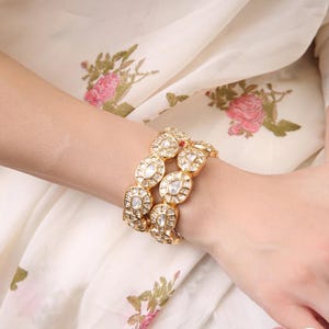 May include: Gold-toned bracelets with clear, round gemstones. The bracelets are worn on a wrist, with a floral patterned fabric in the background. The bracelets have a vintage or antique aesthetic.