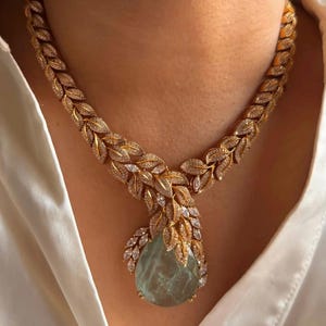 Puede incluir: Un collar de tono dorado con una serie de eslabones entrelazados en forma de hoja incrustados con pequeñas piedras claras. El collar termina en un gran colgante en forma de lágrima de piedra translúcida verde pálido con detalles de hojas.