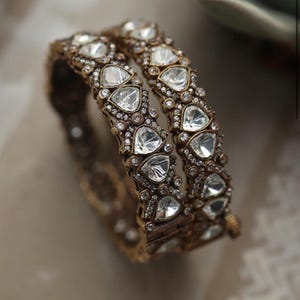 Op de afbeelding: Twee antieke gouden armbanden met heldere, hartvormige stenen en kleinere, ronde accentstenen. De armbanden zijn sierlijk en gedetailleerd, met een vintage esthetiek. De sieraden worden gepresenteerd op een neutrale ondergrond.