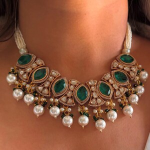 May include: A gold-toned necklace featuring large emerald-green gemstones and small clear stones. The necklace has a pearl strand and dangling pearl accents. The design is ornate and elegant, suitable for special occasions.
