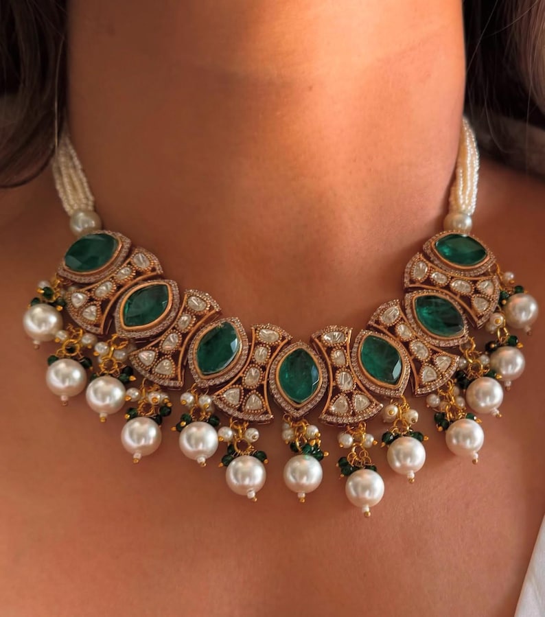 May include: A gold-toned necklace featuring large emerald-green gemstones and small clear stones. The necklace has a pearl strand and dangling pearl accents. The design is ornate and elegant, suitable for special occasions.