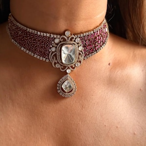 May include: A luxurious choker necklace featuring a central square-cut, clear gemstone pendant, surrounded by a halo of smaller diamonds. The choker band is a deep red with embedded gemstones and a border of small diamonds. A teardrop-shaped pendant hangs below.