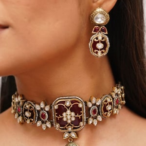 Puede incluir: Un collar gargantilla de oro con un diseño de esmalte rojo y blanco. El collar tiene un colgante que cuelga del centro. El collar está adornado con pequeñas piedras preciosas blancas y rojas.