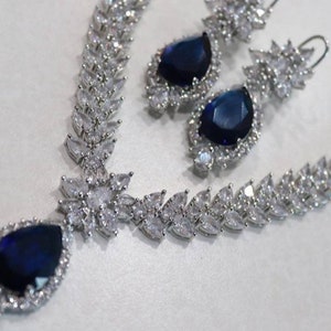 Puede incluir: Un collar y pendientes de plata con un colgante de piedra azul. El collar presenta una cadena delicada con un diseño floral y los pendientes tienen un diseño similar con una piedra azul más pequeña.