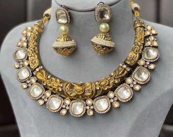 Op de afbeelding: Een gouden en zwarte ketting met een grote, sierlijke hanger en bijpassende oorbellen. De hanger is versierd met ingewikkelde bloemmotieven en heeft een grote, heldere steen in het midden. De oorbellen zijn ook versierd met heldere stenen.