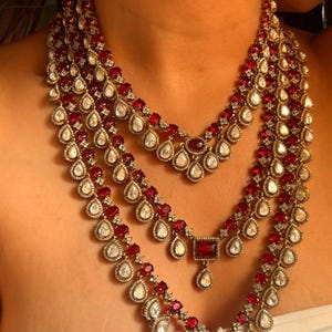 Pode incluir: Colar multicamadas com pedras preciosas vermelhas e transparentes em metal dourado. O colar tem um pendente floral e várias formas, incluindo gotas e quadrados. Joia para usar ao redor do pescoço.