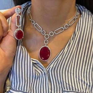 Puede incluir: Un conjunto de collar y pendientes plateados con grandes piedras preciosas ovaladas de color rojo intenso. El collar tiene un colgante y una cadena con un diseño geométrico. Los pendientes tienen un diseño similar. La joyería está engastada con pequeñas piedras transparentes.