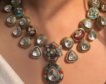 May include: A gold necklace with a teardrop-shaped pendant featuring a large clear stone surrounded by smaller colorful stones. The necklace is made up of multiple smaller teardrop-shaped stones with gold accents.