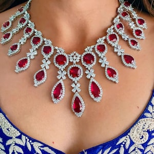 Peut inclure: Un collier éblouissant orné de grandes pierres précieuses rouges de forme ovale et en goutte, serties dans un métal argenté. Le collier présente un motif floral avec de plus petites pierres claires entourant les gemmes rouges. Le collier est porté sur un vêtement bleu.