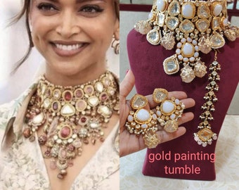 May include: A gold necklace set with white and pink stones, featuring a large choker necklace, matching earrings, and a long pendant necklace. The set is displayed on a red velvet stand with the text "gold painting tumble".