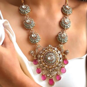 May include: A gold necklace with a large, ornate pendant featuring a circular design with white and light blue stones. The pendant is surrounded by smaller, teardrop-shaped pink stones.