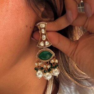 May include: Gold-toned earring with a geometric top section, featuring clear and red stones. The earring has a large, oval, emerald-green stone at the center, surrounded by smaller clear stones. Below, it dangles with pearls and green beads. The earring is being held up to an ear.