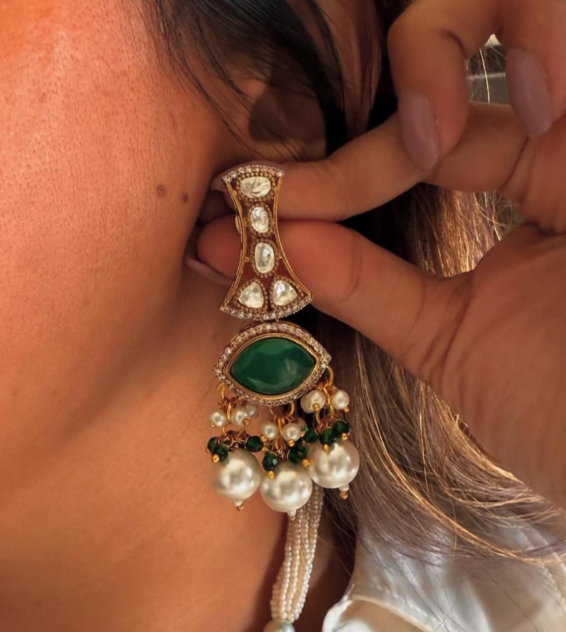 May include: Gold-toned earring with a geometric top section, featuring clear and red stones. The earring has a large, oval, emerald-green stone at the center, surrounded by smaller clear stones. Below, it dangles with pearls and green beads. The earring is being held up to an ear.