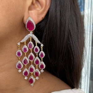Könnte beinhalten: Elegante Chandelier-Ohrringe mit tropfenförmigen rubinroten Edelsteinen. Die Ohrringe haben eine goldfarbene Fassung mit klaren, funkelnden Akzenten. Das Design umfasst einen großen Rubin oben und kaskadierende Edelsteine darunter.