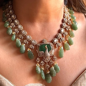 Puede incluir: Un collar de oro con un gran colgante verde esmeralda con un diseño de pavo real. El colgante está rodeado de diamantes más pequeños y el collar está adornado con piedras verdes en forma de lágrima.